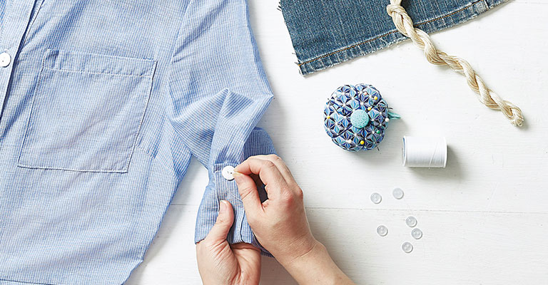 hands sewing a white button to a blue blouse next to sewing supplies, a pair of blue jeans, and a blue blouse
